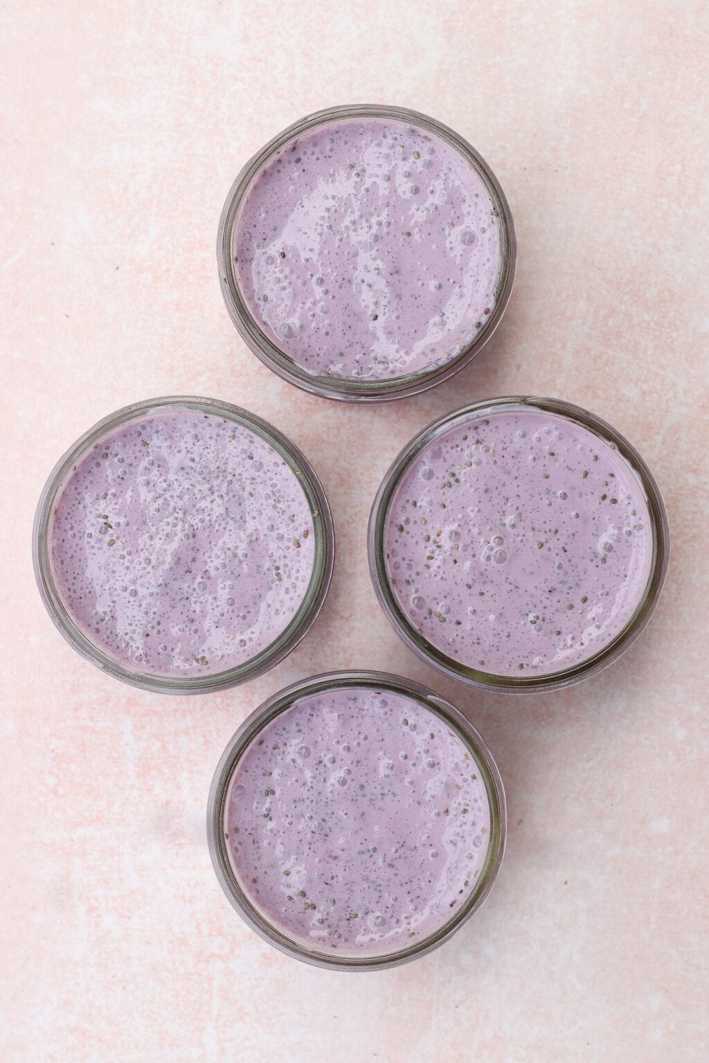 Four glass jars filled with creamy purple smoothies are arranged on a light pink textured surface.