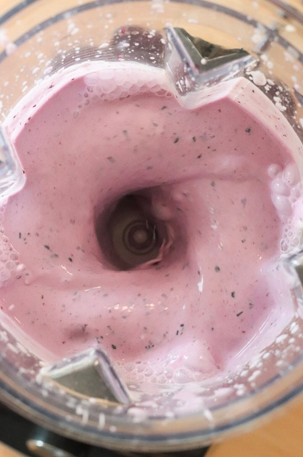 Close-up view of a blender mixing a creamy, light pink smoothie with small dark specks, creating a swirling vortex in the center.