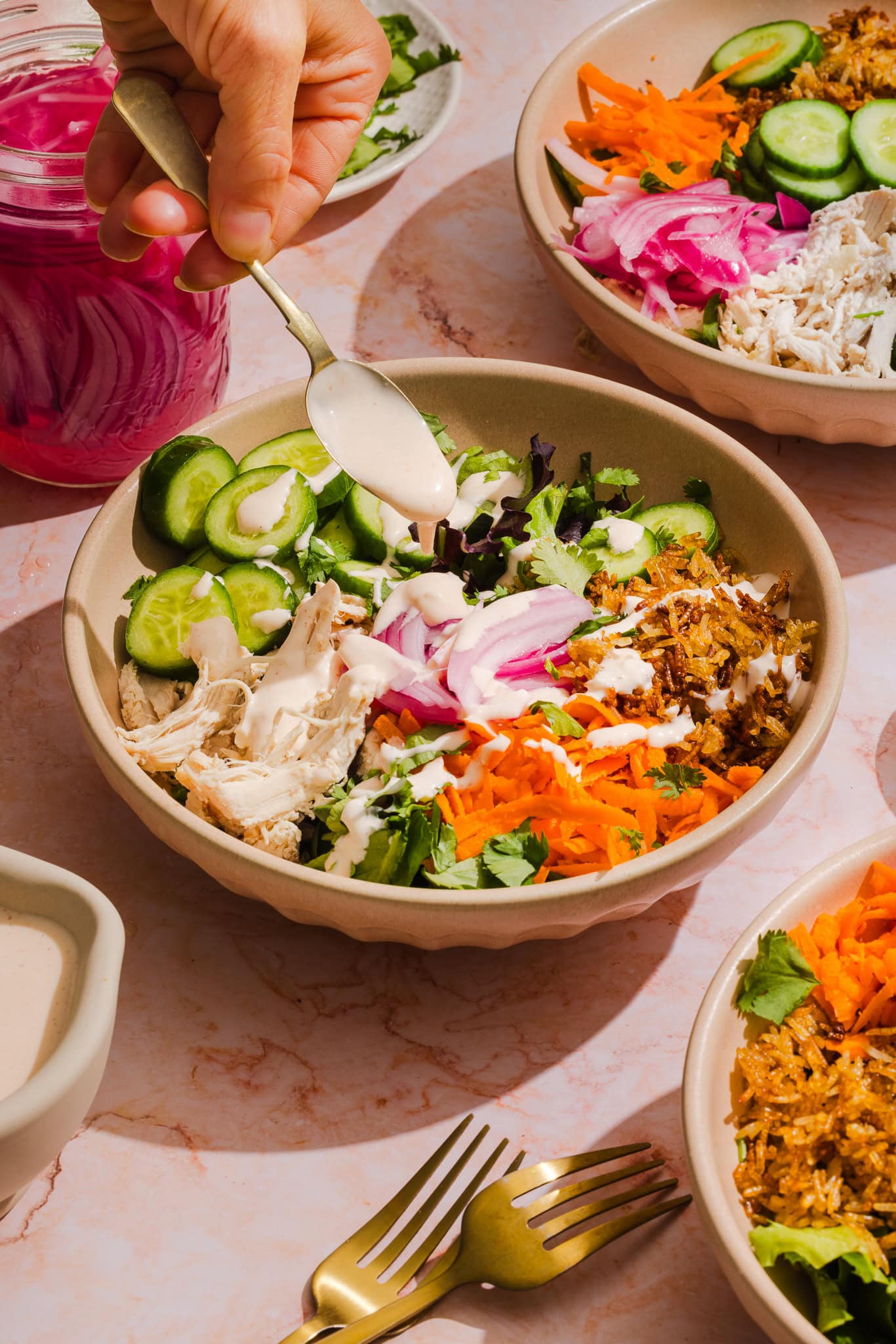 A hand drizzles creamy white dressing over a bowl filled with shredded chicken, pickled onions, cucumbers, carrots, greens, and cilantro. Other similar bowls and a jar of pickled onions are on a light pink surface.