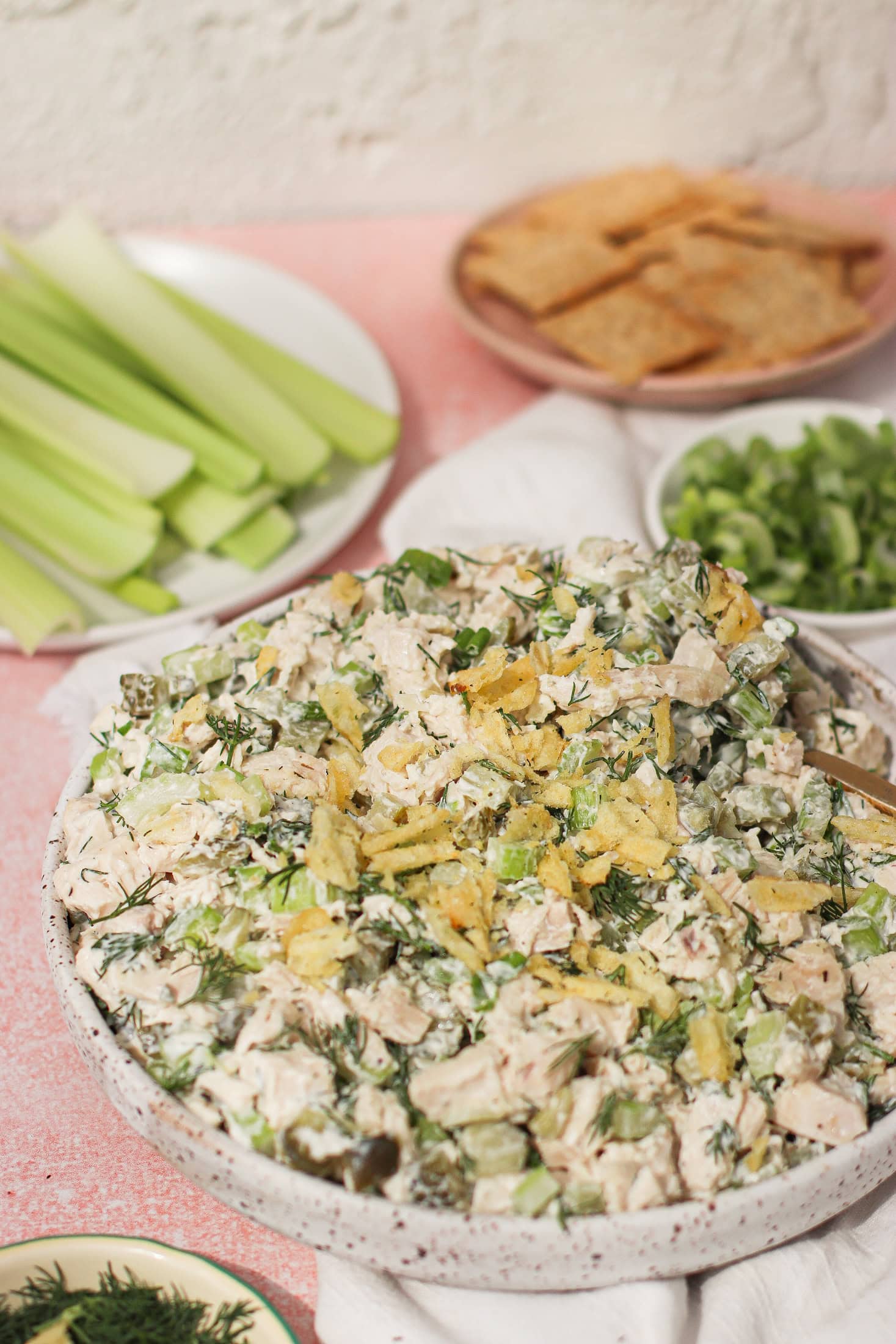 A bowl of creamy chicken salad garnished with crumbled potato chips sits on a table, surrounded by plates of celery sticks, crackers, and sliced green onions.