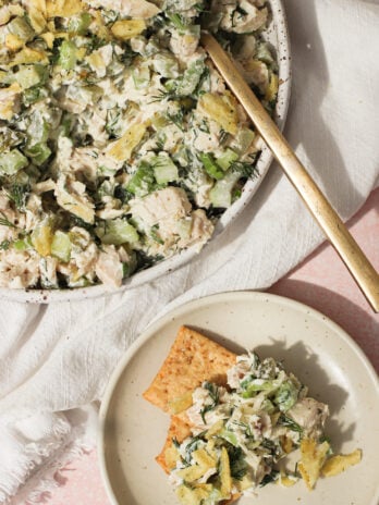 A bowl of chicken salad with celery, herbs, and crushed potato chips, served with a gold fork. A small plate nearby holds crackers topped with the same chicken salad mixture.