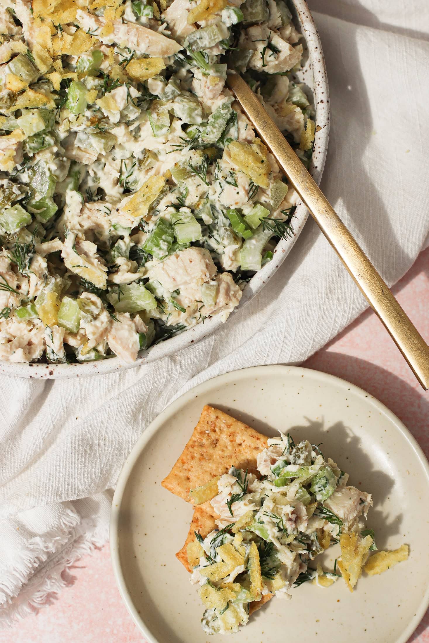 A bowl of chicken salad with celery, herbs, and crushed potato chips, served with a gold fork. A small plate nearby holds crackers topped with the same chicken salad mixture.