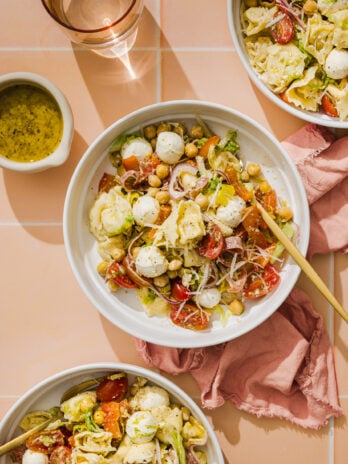 Three bowls of pasta salad with mozzarella balls, chickpeas, cherry tomatoes, lettuce, and shredded cheese on a peach tiled surface, next to a cup of water, a small dish of dressing, and a pink napkin.
