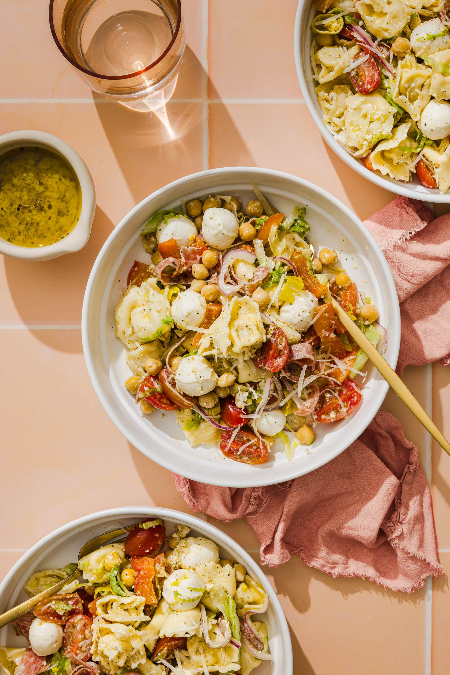 Three bowls of pasta salad with mozzarella balls, chickpeas, cherry tomatoes, lettuce, and shredded cheese on a peach tiled surface, next to a cup of water, a small dish of dressing, and a pink napkin.