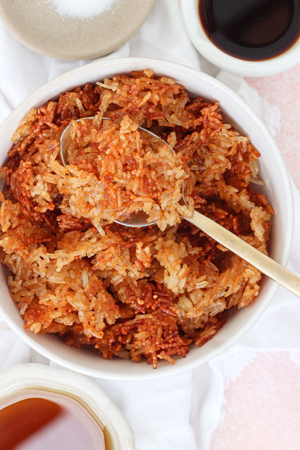 A bowl of crispy, browned fried rice with a spoon scooping some out. Small dishes of soy sauce and salt are placed nearby on a white cloth.