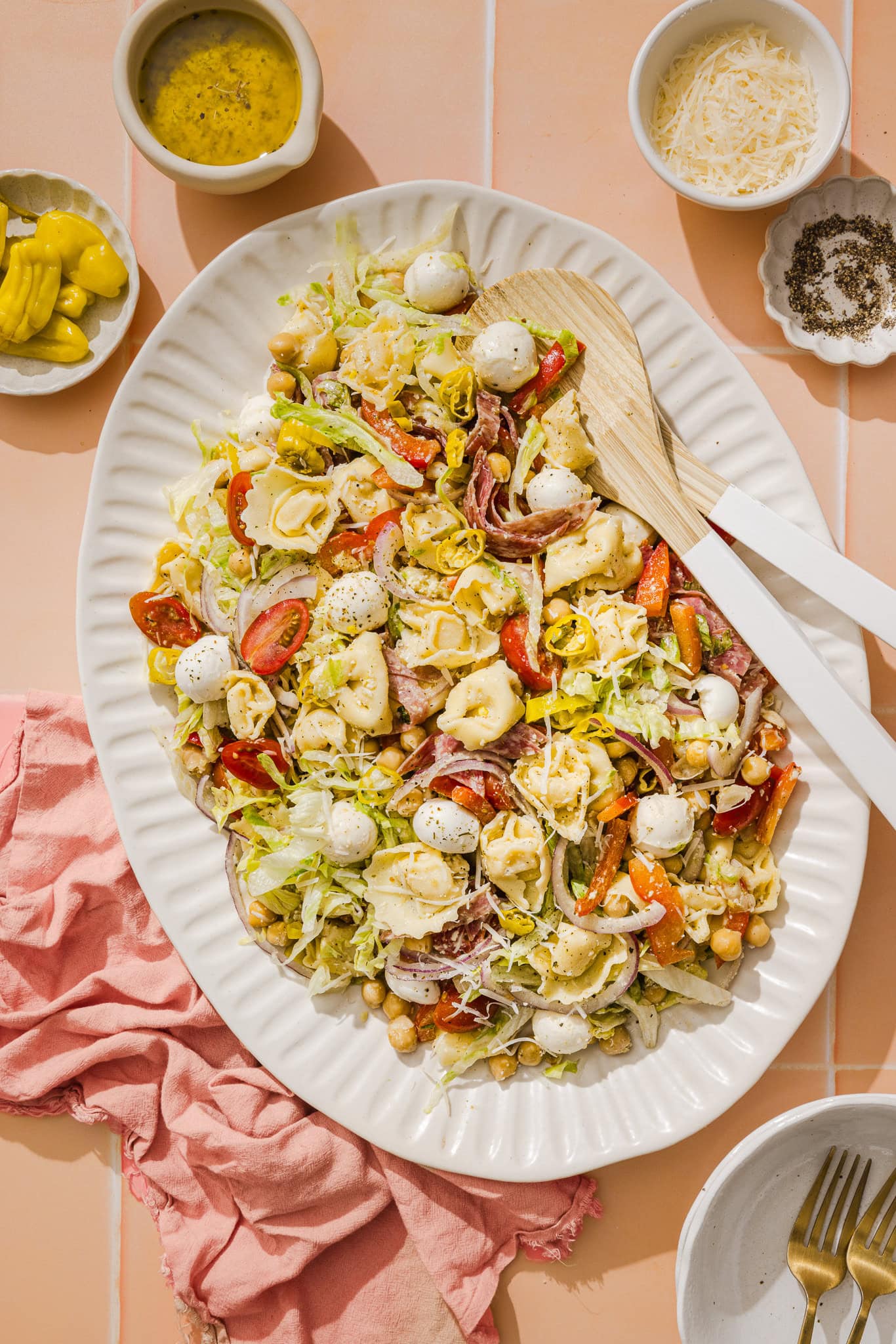 A large oval platter of Italian chopped salad with tortellini, mozzarella balls, cherry tomatoes, lettuce, chickpeas, and peppers—like a vibrant grinder pasta salad—surrounded by bowls of dressing, parmesan, pepper, and a pink napkin on peach tile.