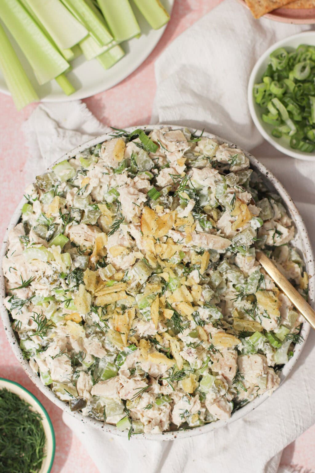 A bowl of chicken salad mixed with herbs and topped with crushed potato chips, surrounded by celery sticks, sliced green onions, and dill on a light pink surface.