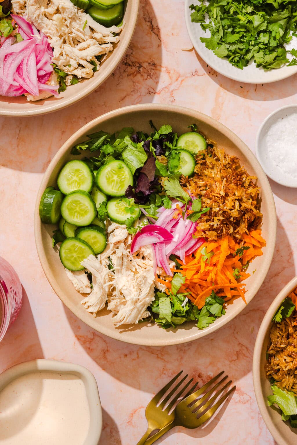 A colorful salad bowl with shredded chicken, sliced cucumbers, pickled onions, shredded carrots, greens, cilantro, and crispy fried onions, surrounded by plates of herbs, salt, a sauce, and two forks on a marble surface.