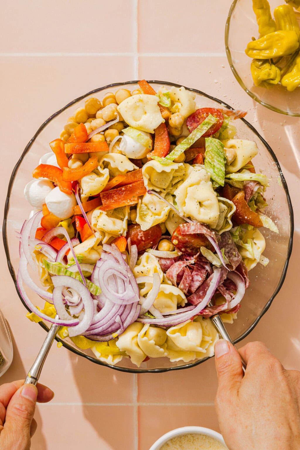 A person tosses a large bowl of pasta salad with tortellini, sliced red onions, bell peppers, salami, chickpeas, mozzarella balls, celery, and carrots, using two serving spoons.