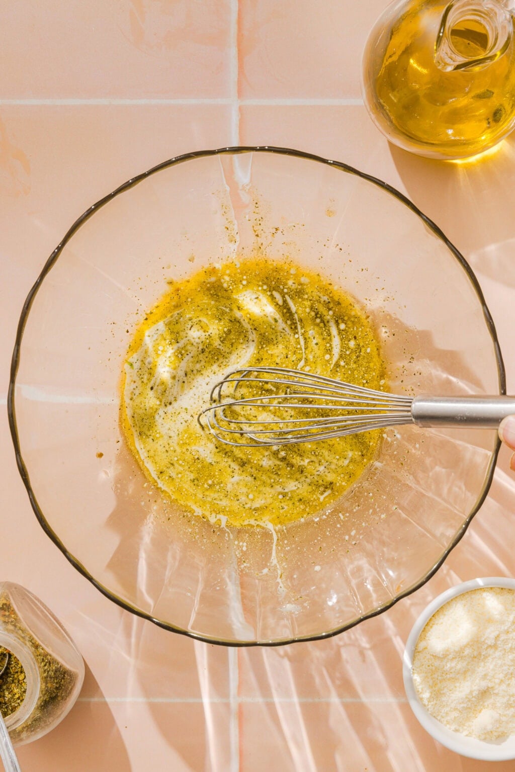 A hand uses a metal whisk to mix oil and seasonings in a glass bowl on a light tiled surface, with jars of spices, grated cheese, and a bottle of oil nearby.