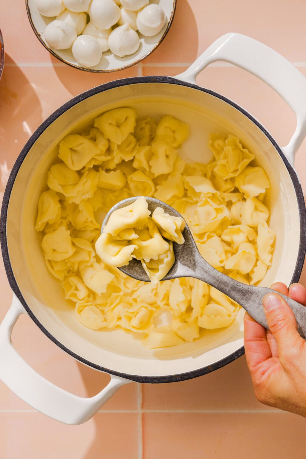 A hand holds a large spoon, scooping cooked cheese tortellini from a white pot. In the background, a plate with small mozzarella balls is visible on a light pink tiled surface.