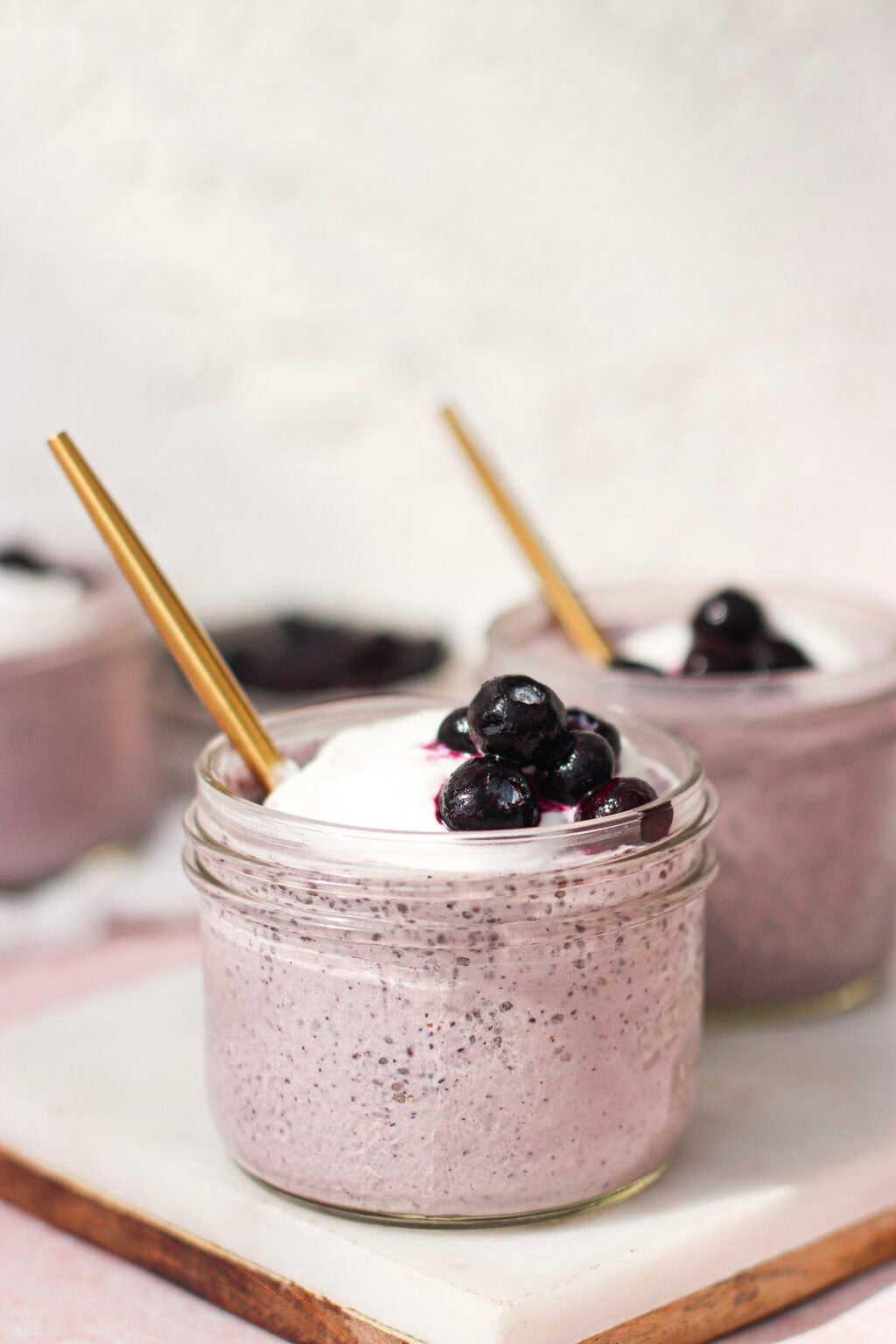 A glass jar filled with a creamy purple smoothie, topped with whipped cream and blueberries, with a gold straw. Another similar jar is in the background, both placed on a white surface.
