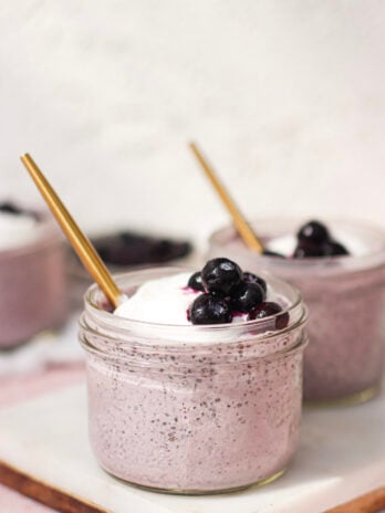 A glass jar filled with a creamy purple smoothie, topped with whipped cream and blueberries, with a gold straw. Another similar jar is in the background, both placed on a white surface.