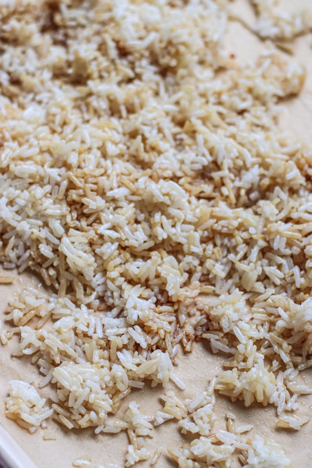 Cooked white rice spread out on a flat surface, with some grains showing a light brown, toasted color. The rice appears separated and slightly uneven in texture.
