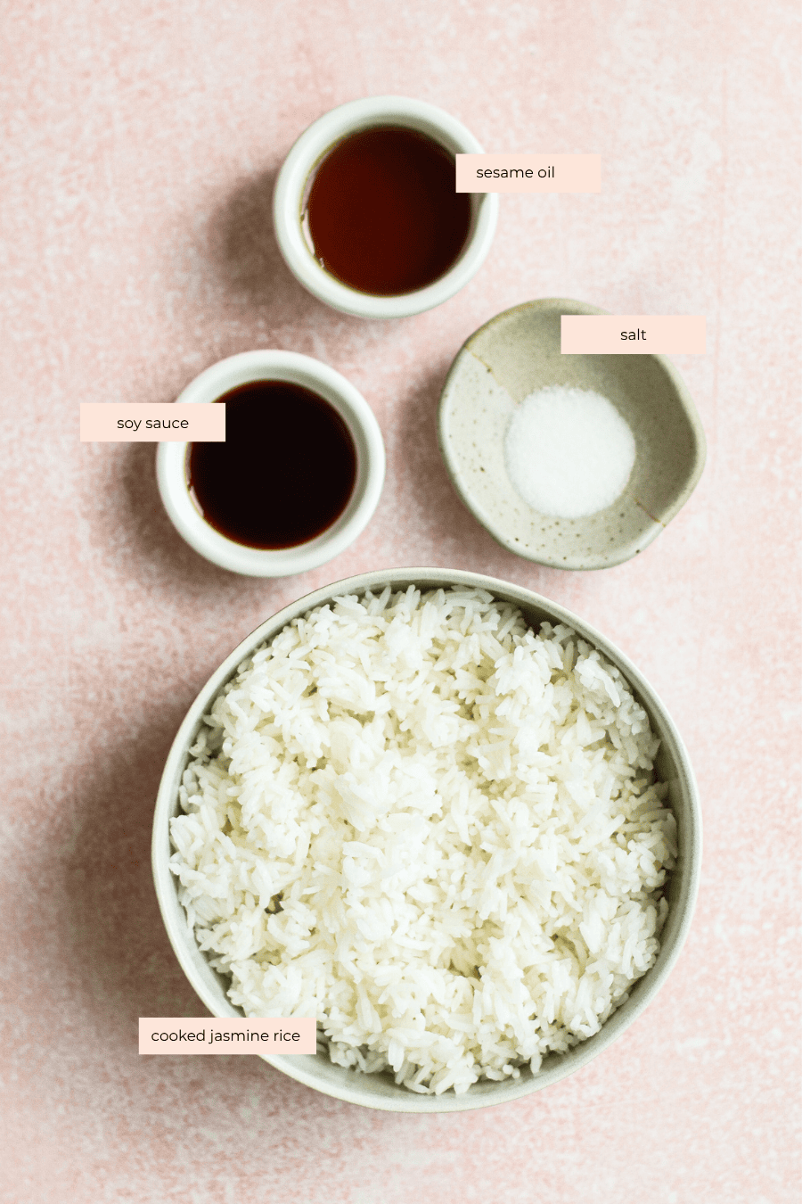 A bowl of cooked jasmine rice with small bowls containing soy sauce, sesame oil, and salt arranged on a light pink surface. Each ingredient is labeled with text.