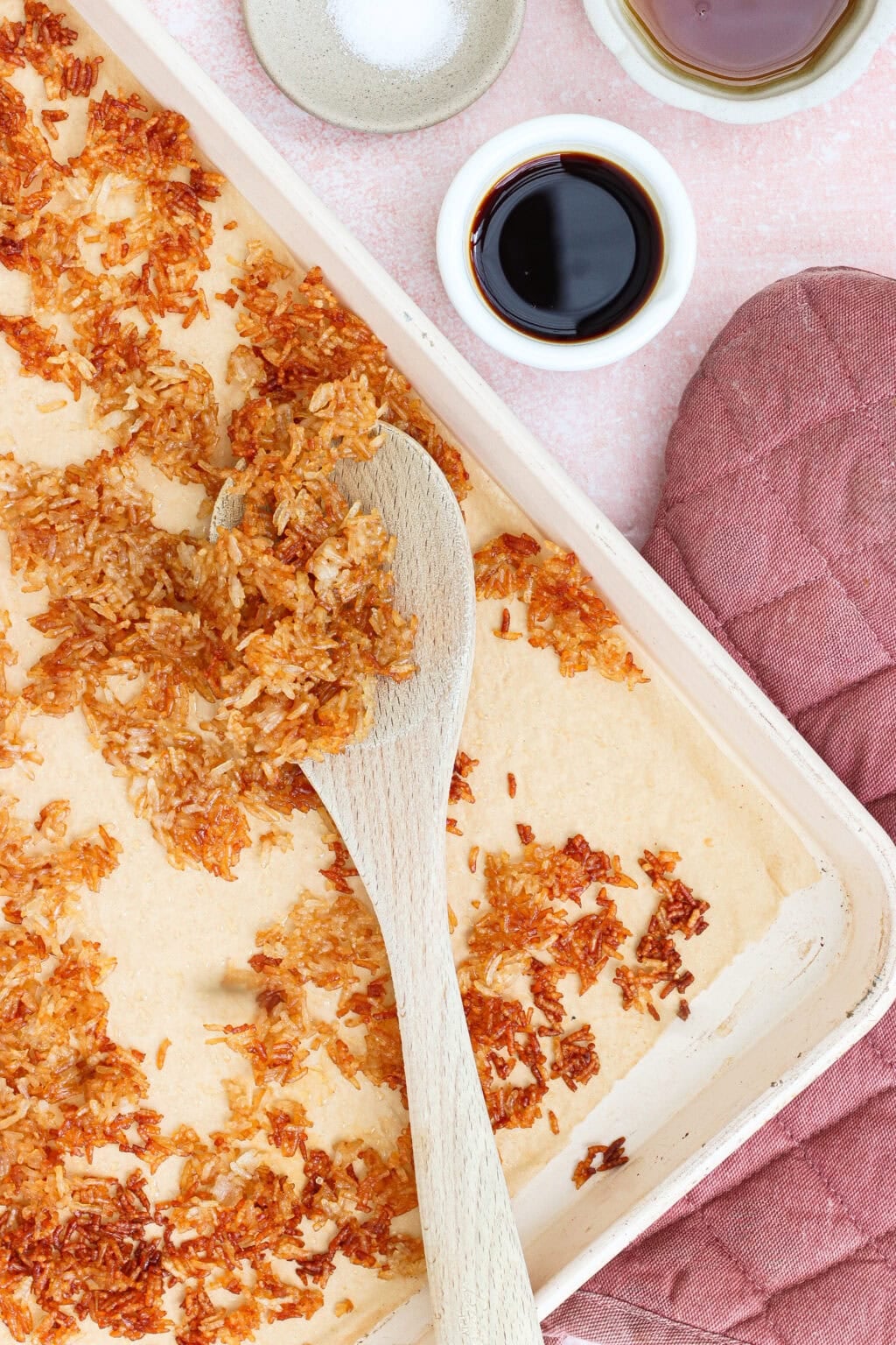 A baking tray with toasted coconut flakes being stirred with a wooden spoon, surrounded by a pink oven mitt, a bowl of salt, and two small bowls with dark liquid.