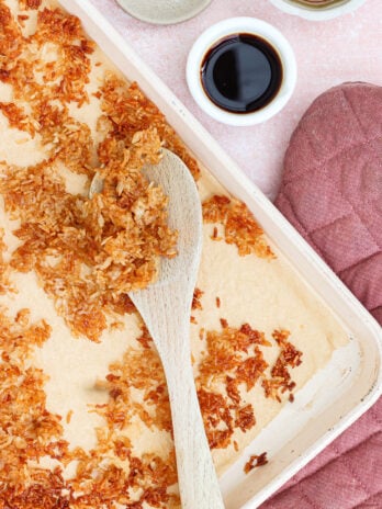 A baking tray with toasted coconut flakes being stirred with a wooden spoon, surrounded by a pink oven mitt, a bowl of salt, and two small bowls with dark liquid.