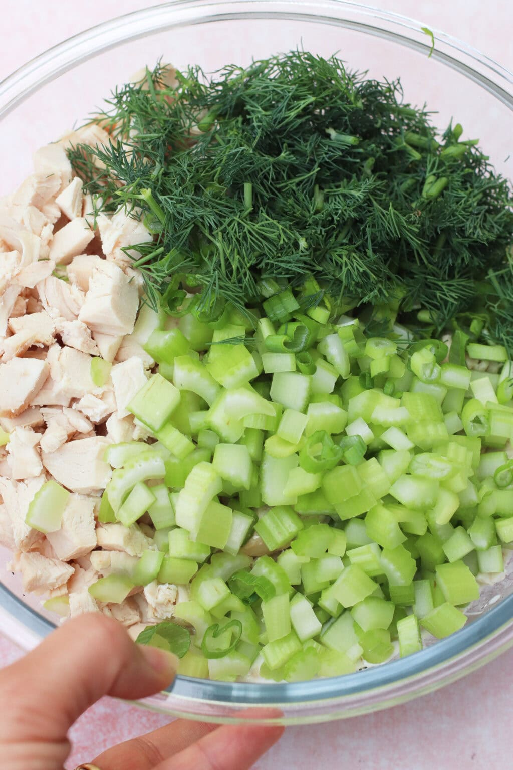 A clear glass bowl containing chopped cooked chicken, fresh dill, chopped celery, and sliced green onions, with a hand holding the side of the bowl.