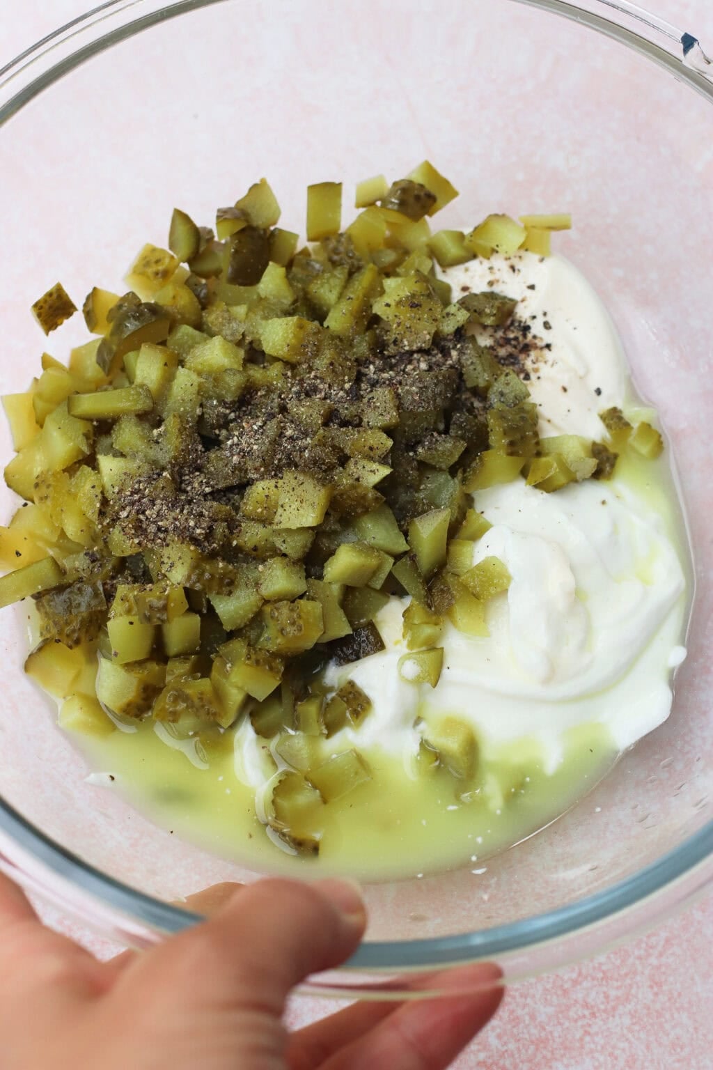 A clear glass bowl containing chopped pickles, yogurt or sour cream, pickle juice, and ground black pepper, held by a hand against a light pink background.