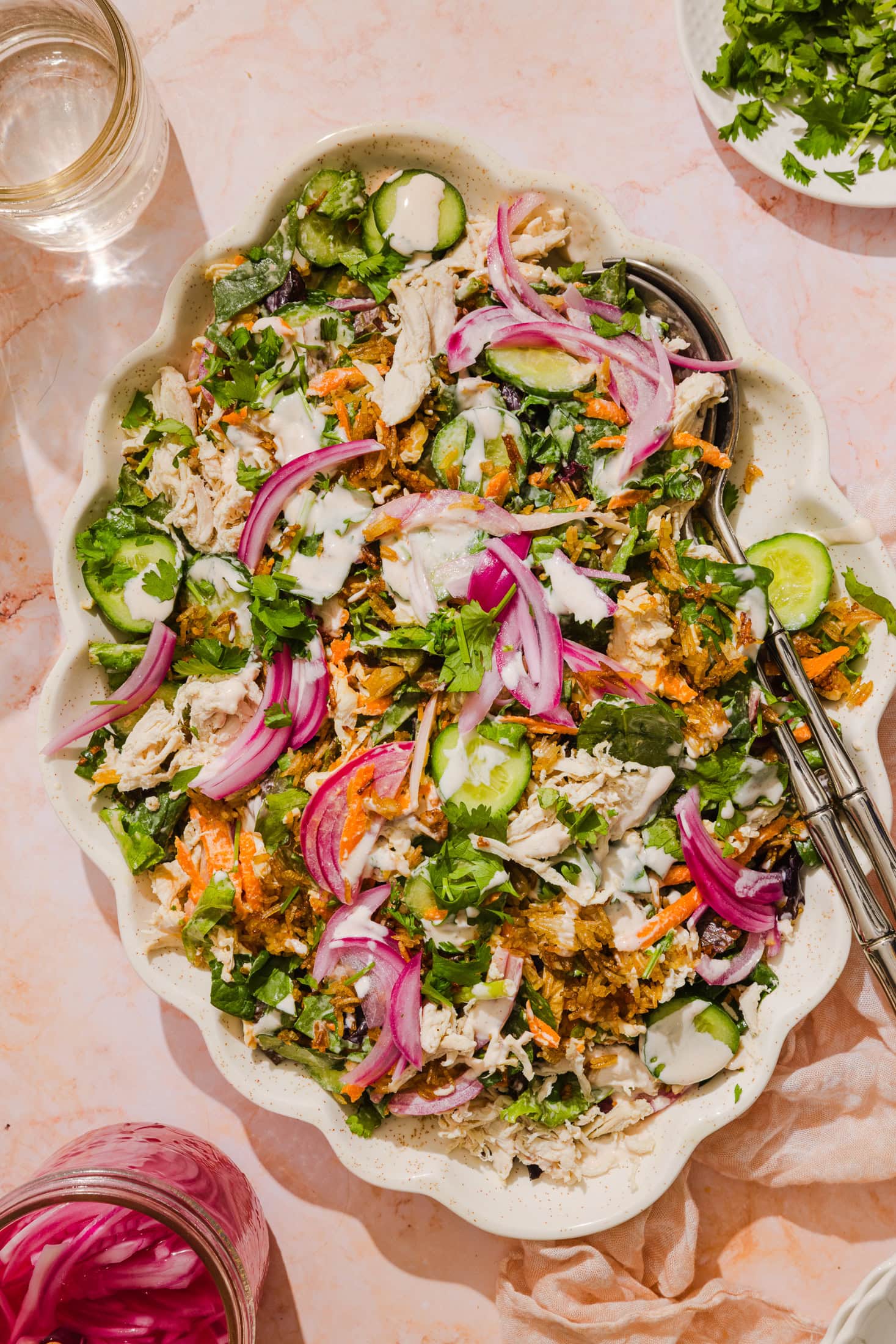 A large scalloped bowl filled with a colorful salad of shredded chicken, sliced cucumber, pickled red onions, grated carrots, chopped greens, and fresh cilantro, drizzled with a creamy dressing. Tongs rest on the side of the bowl.