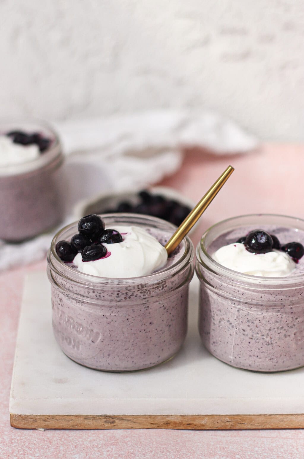 Two small glass jars filled with creamy purple blueberry chia pudding smoothie, topped with whipped cream and blueberries. One jar has a gold spoon inside. The jars sit on a white board with a light pink background.