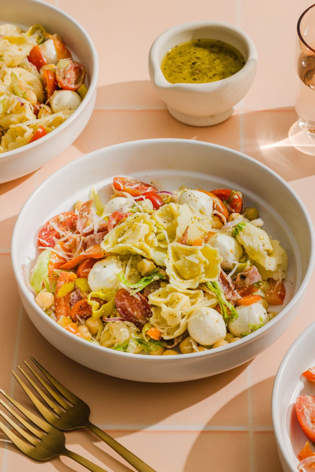 A bowl of tortellini pasta salad with mozzarella balls, cherry tomatoes, lettuce, and sliced salami, served with a side of green dressing. Gold forks and other salad bowls are visible on a peach-colored tiled surface.