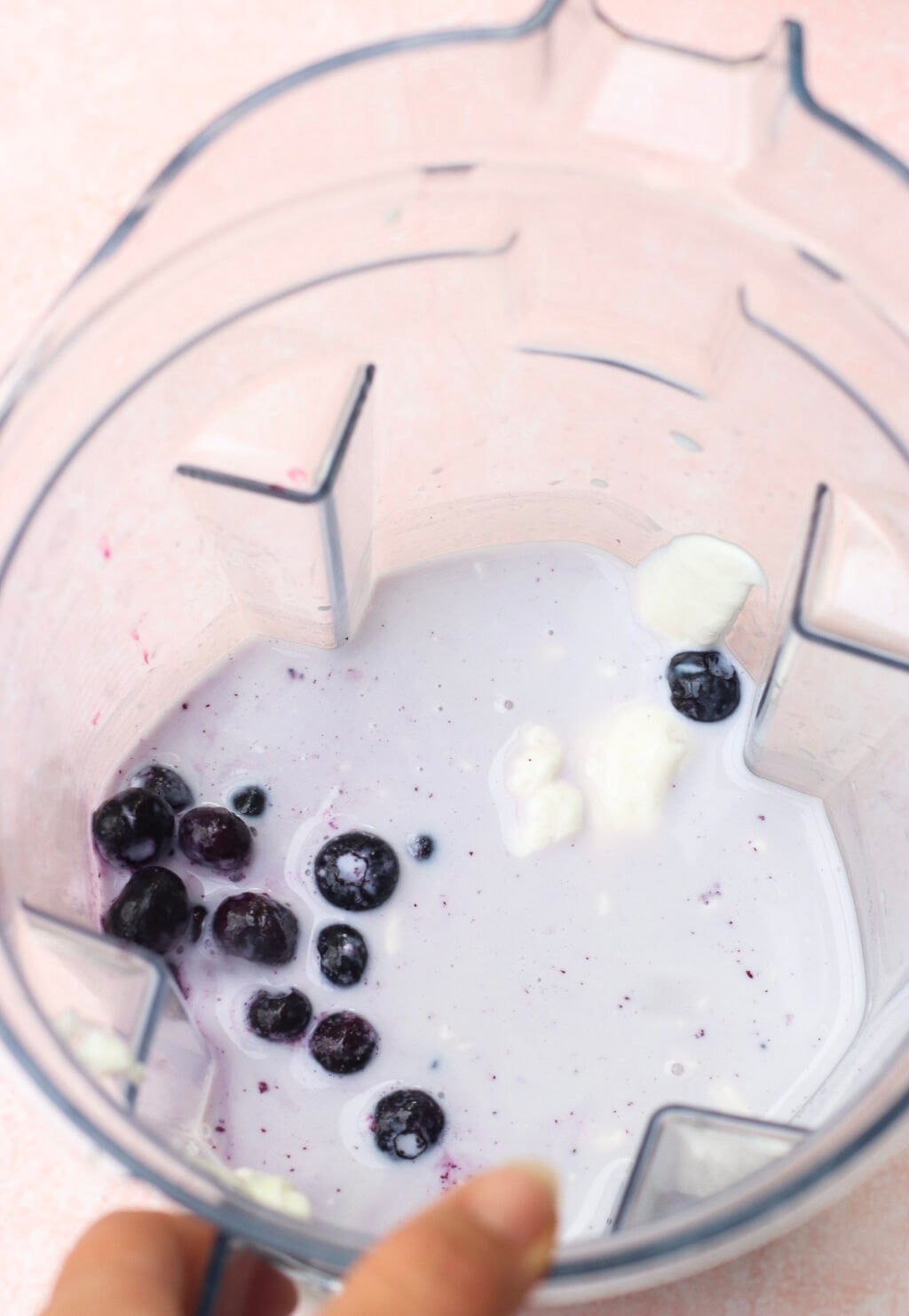 A top-down view of a blender containing a small amount of milk or yogurt, several blueberries, and a few dollops of yogurt. A hand is holding the edge of the blender.