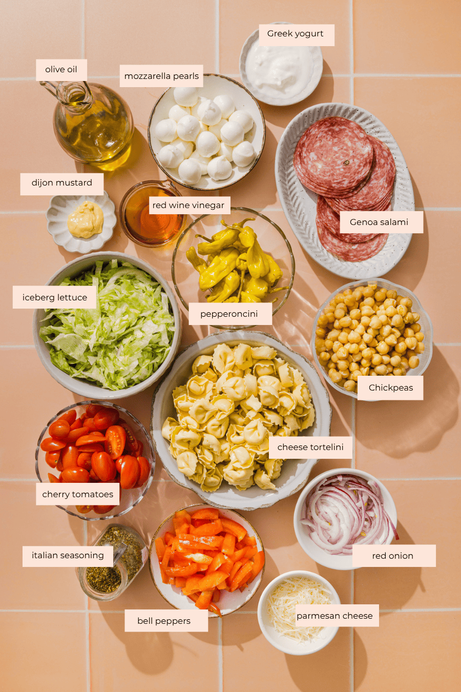 A colorful spread of ingredients in bowls on a tiled surface, including olive oil, mozzarella pearls, Greek yogurt, Genoa salami, lettuce, pepperoncini, chickpeas, tortellini, tomatoes, bell peppers, onion, and cheese.