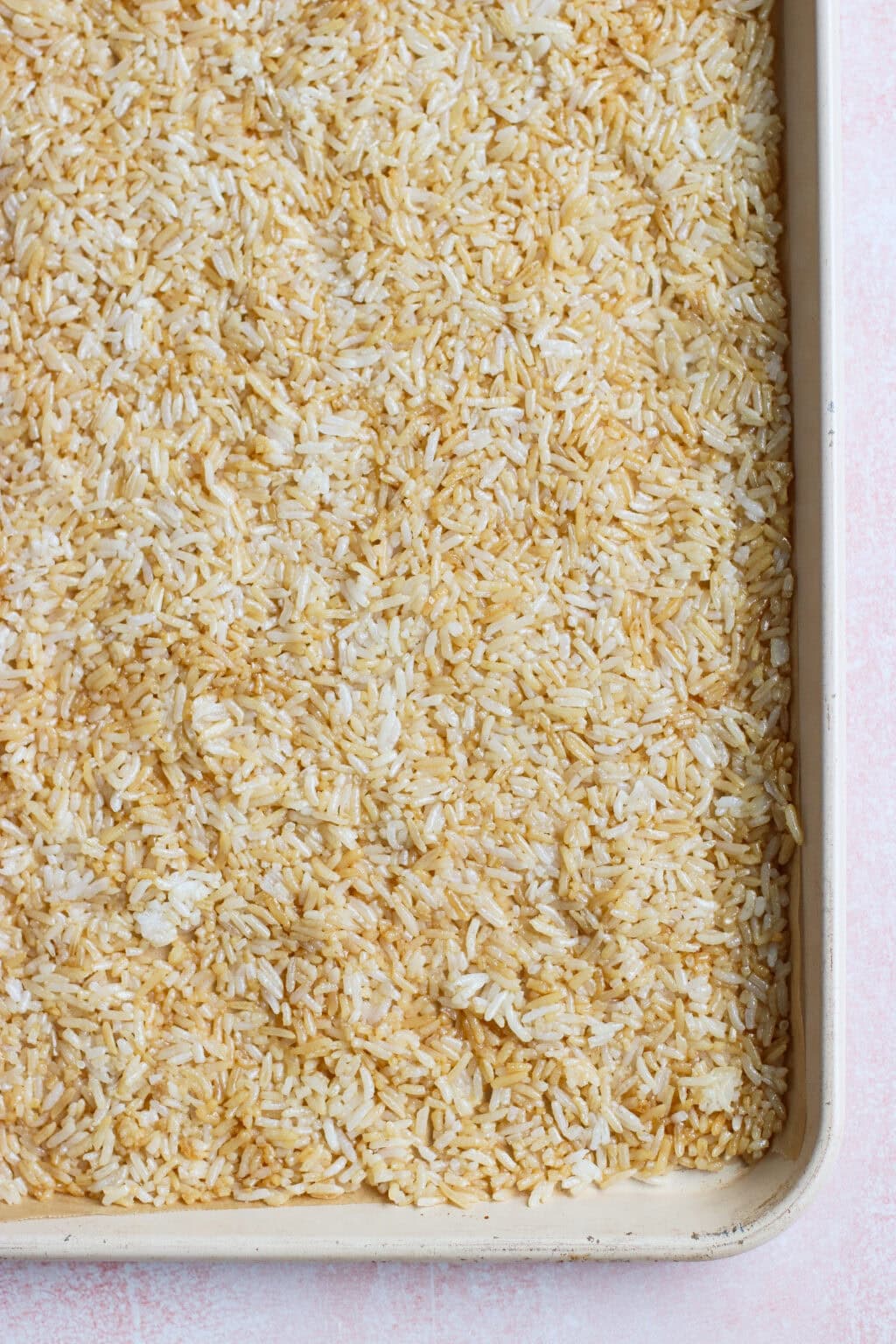 Uncooked white rice spread evenly on a large rimmed baking sheet, showing a mix of light and slightly toasted grains.