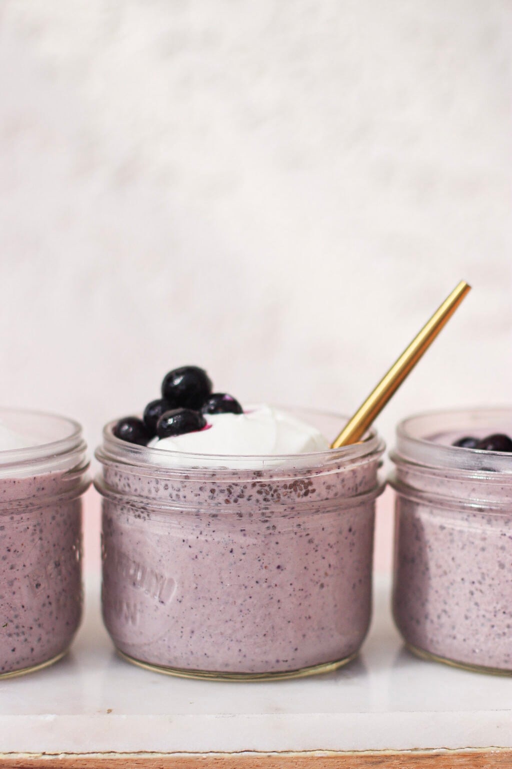 Three glass jars filled with creamy purple smoothie, reminiscent of blueberry chia pudding, topped with whipped cream and blueberries. The center jar has a gold spoon inserted. The jars are arranged in a row against a light background.