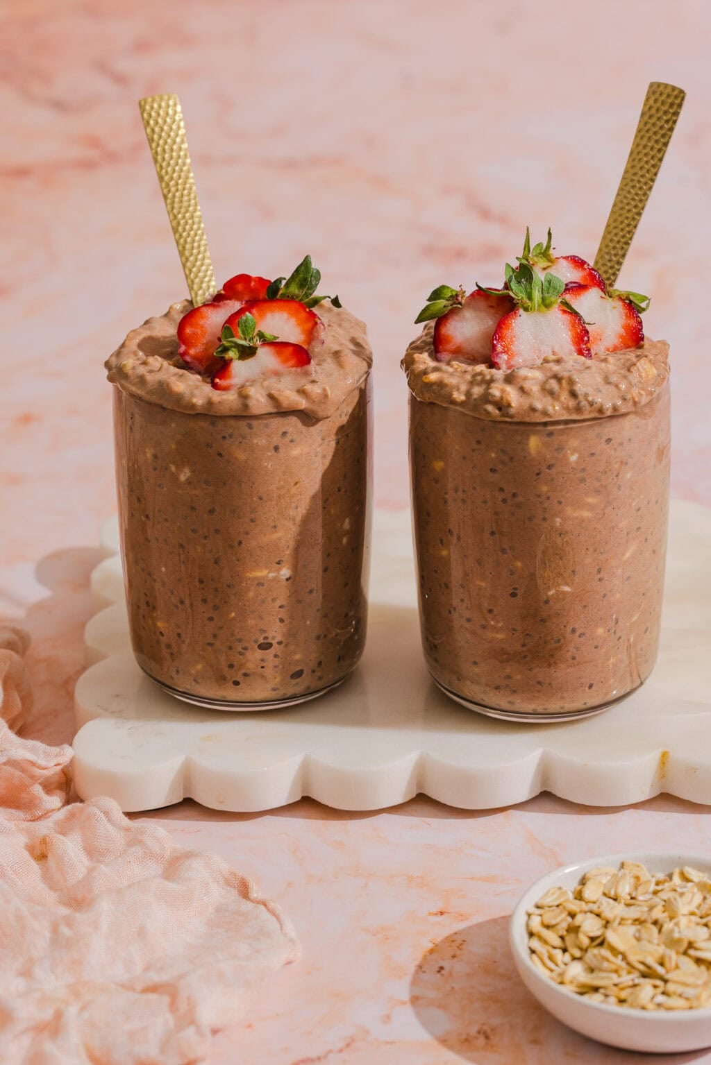 Two glasses of chocolate overnight oats topped with sliced strawberries and gold spoons, placed on a white tray, with a small bowl of oats nearby on a pink surface.