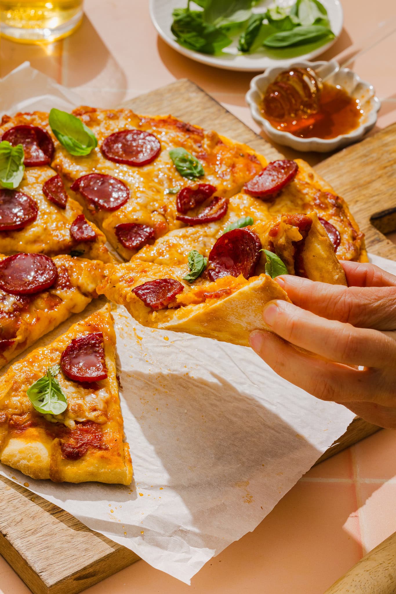 A hand lifts a slice of pepperoni pizza from a wooden board, with melted cheese and basil leaves on top. A bowl of fresh basil and a dish of honey are in the background.