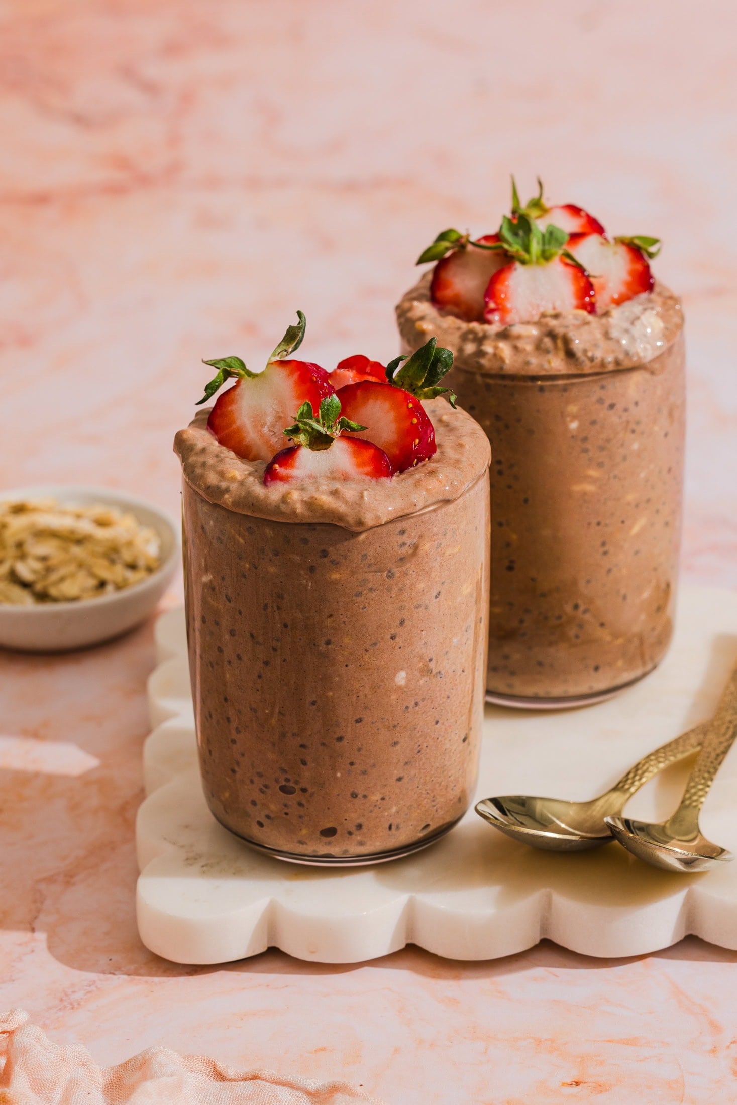 Two glasses of chocolate chia pudding topped with sliced strawberries sit on a white tray, with two gold spoons and a small bowl of nuts beside them, all on a pink marble surface.