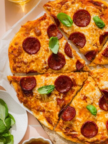 Pepperoni pizza sliced on a wooden board, topped with fresh basil leaves. A glass of drink and a bowl of fresh basil leaves are nearby.