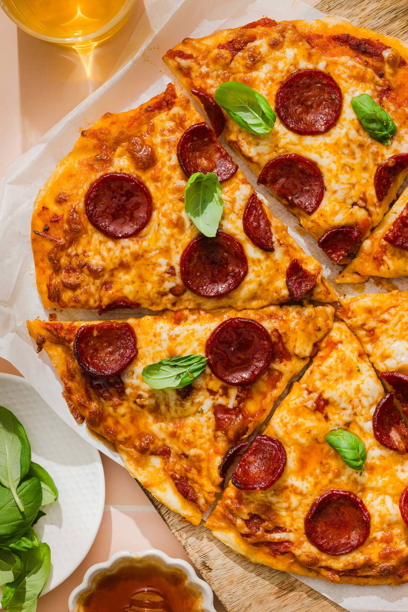 Pepperoni pizza sliced on a wooden board, topped with fresh basil leaves. A glass of drink and a bowl of fresh basil leaves are nearby.