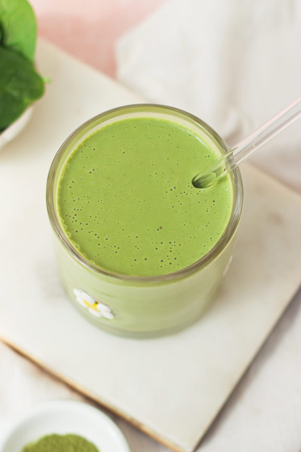 A glass filled with a creamy green smoothie, topped with tiny bubbles, has a clear straw and sits on a white surface. A small white flower is on the outside of the glass.
