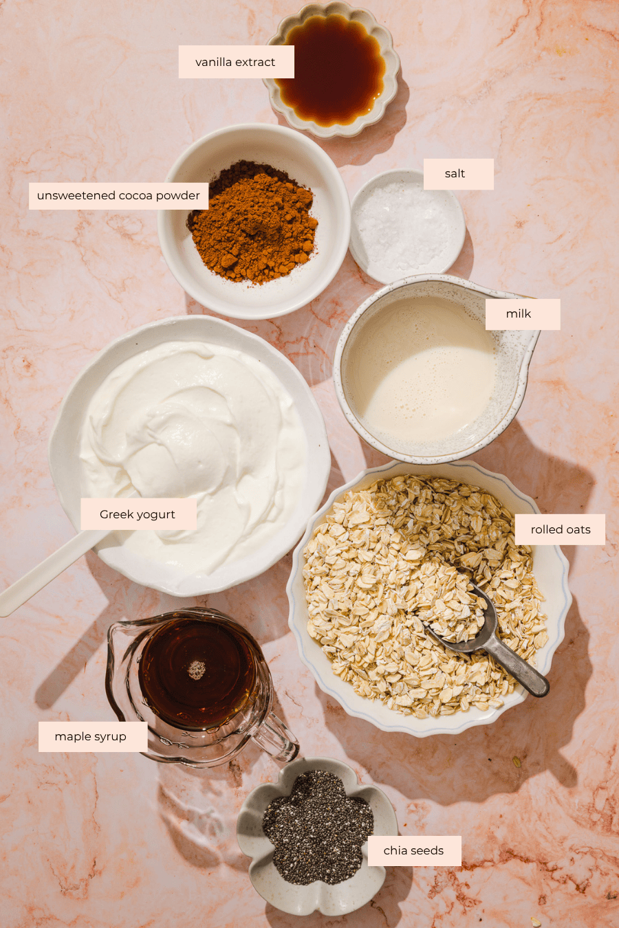 Bowls of Greek yogurt, unsweetened cocoa powder, salt, milk, rolled oats, maple syrup, chia seeds, and vanilla extract are arranged on a marble surface, each ingredient labeled with text.