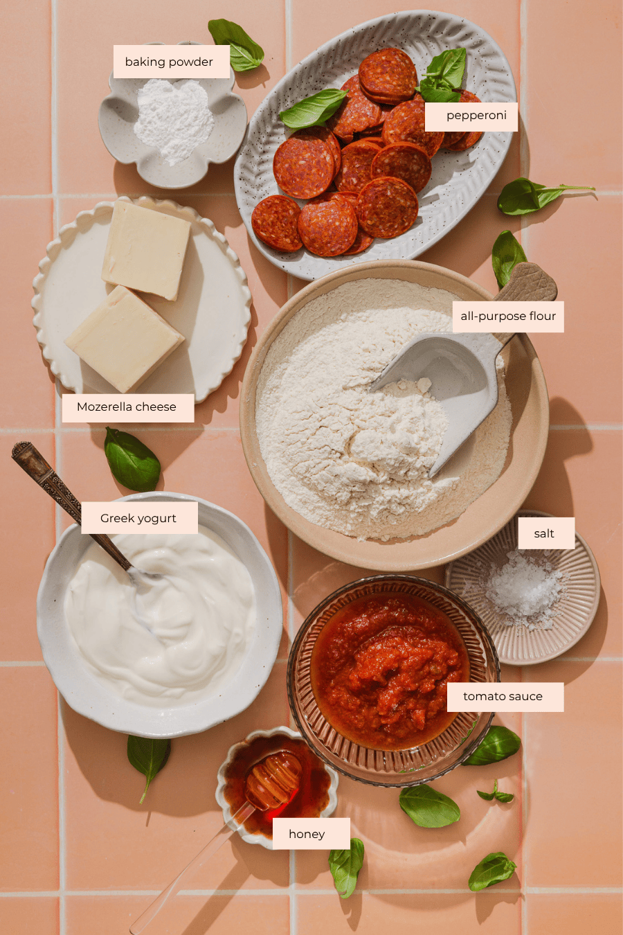 Bowls of flour, baking powder, salt, Greek yogurt, tomato sauce, honey, mozzarella cheese, and pepperoni slices are arranged neatly on a pink tile surface, garnished with fresh basil leaves.