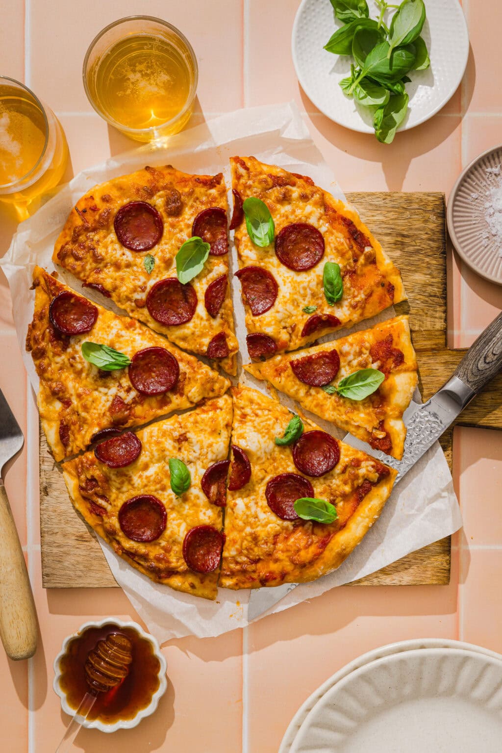 A sliced pepperoni pizza with fresh basil on a wooden board, surrounded by two drinks, a knife, basil leaves, grated cheese, and a small bowl of honey on a peach-colored tiled surface.