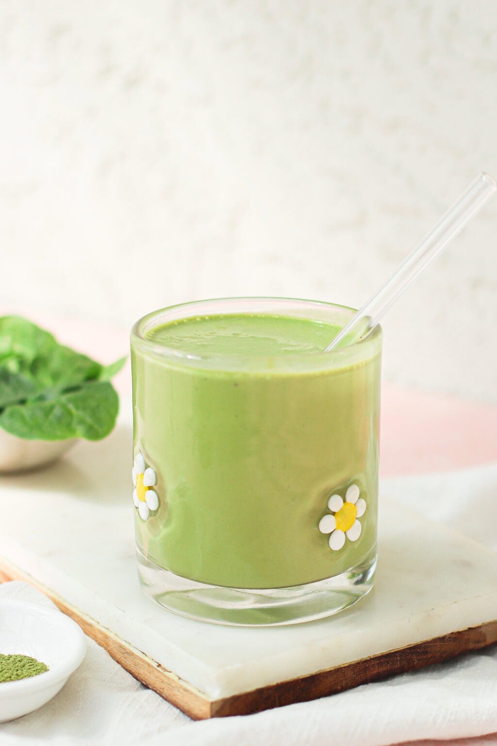 A green smoothie in a glass decorated with daisy flowers, with a clear straw. Spinach leaves and a small dish with green powder are nearby on a marble tray.