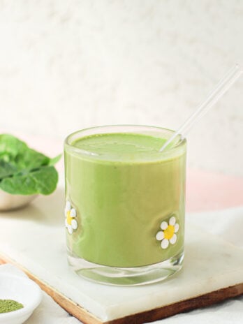 A green smoothie in a glass decorated with daisy flowers, with a clear straw. Spinach leaves and a small dish with green powder are nearby on a marble tray.