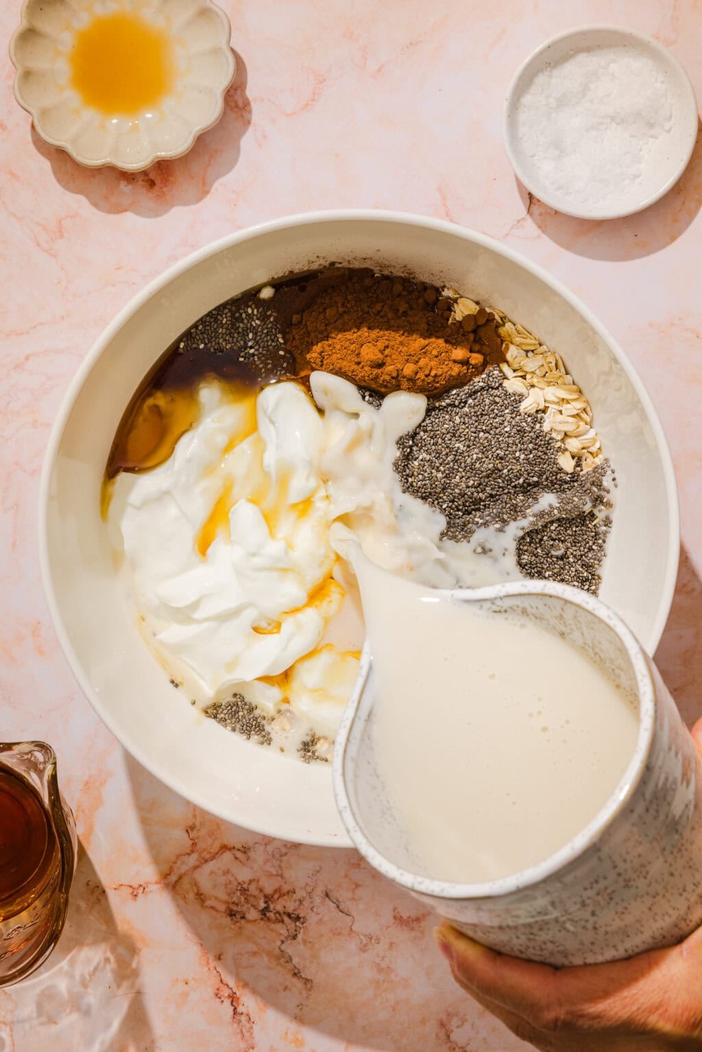 A hand pours milk into a bowl containing yogurt, oats, chia seeds, cinnamon, and honey, with small dishes of honey and salt nearby on a marble surface.