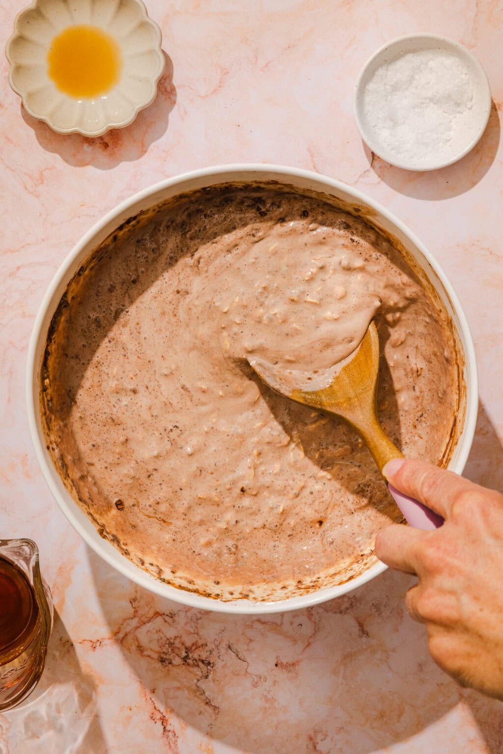 A hand stirs a bowl of chocolate batter with a wooden spoon on a marble surface; small dishes with salt and liquid ingredients are nearby.