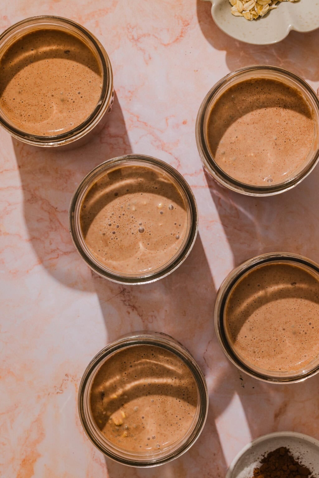 Five glass jars filled with chocolate smoothie are arranged on a light marbled surface. The sunlight casts long shadows from the jars. Some oats and cocoa powder are visible in small bowls nearby.