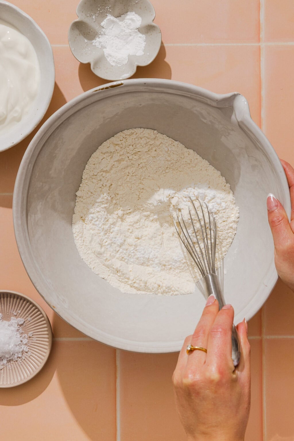 A person whisks flour in a large bowl, surrounded by small dishes of baking powder, salt, and a bowl of yogurt on a peach-colored tiled surface.