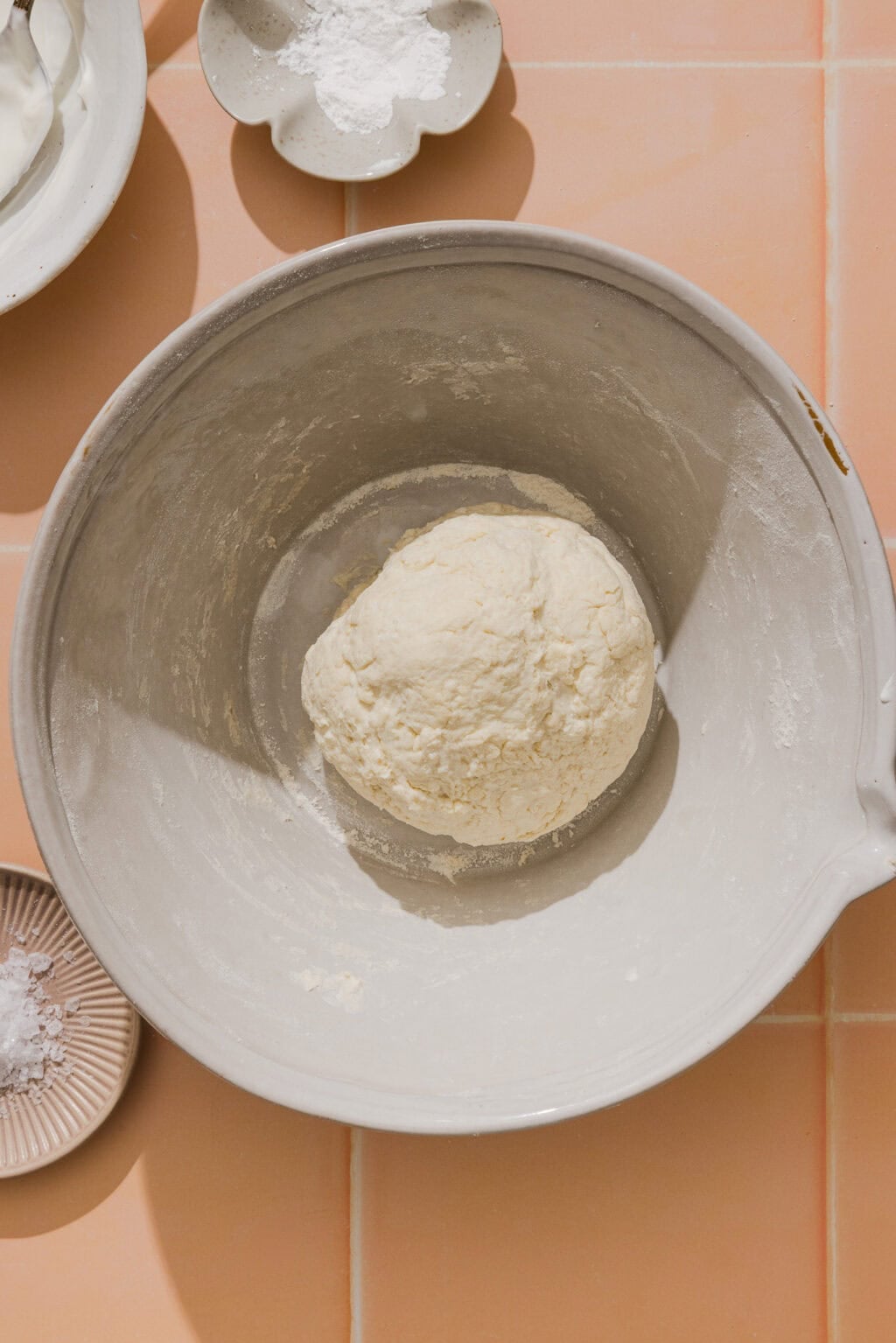 A ball of dough sits in a large mixing bowl on a peach-colored tiled surface, surrounded by small dishes containing flour and other baking ingredients.