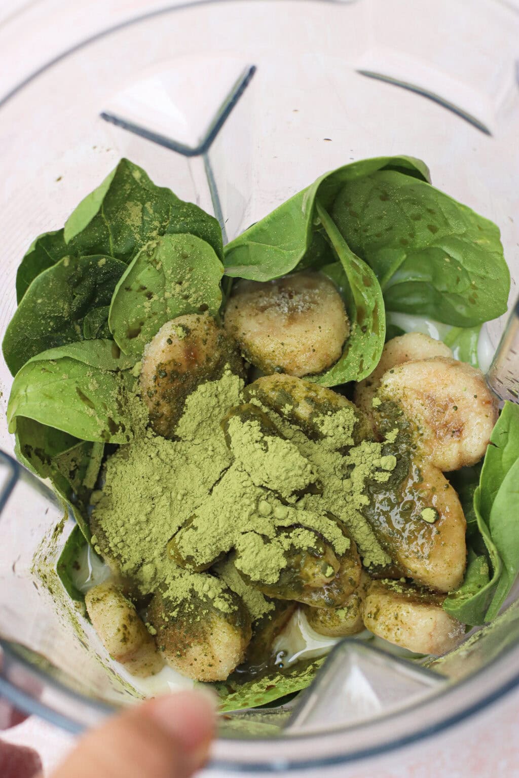 Fresh spinach leaves, banana slices, green powder (likely matcha), and liquid are in a blender, ready to be mixed for a smoothie.