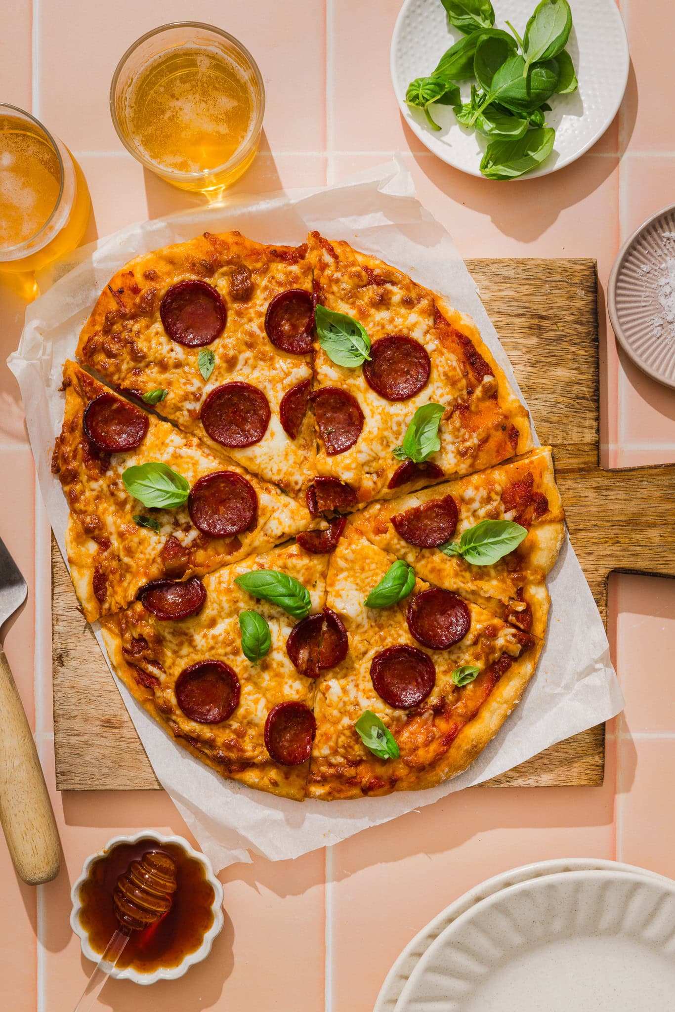 A pepperoni pizza made with greek yogurt pizza dough and topped with fresh basil sits on a wooden board, surrounded by drinks, fresh basil leaves, a small bowl of honey, and empty plates on a pink tiled surface.
