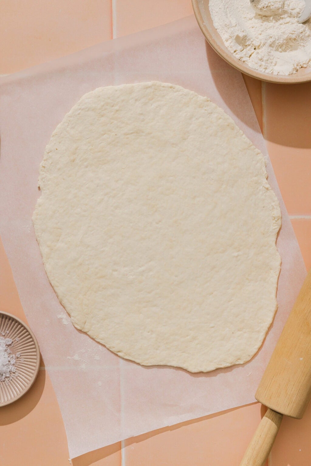 Rolled out pizza dough on parchment paper, surrounded by a bowl of flour, a rolling pin, and a small dish of salt on a peach-colored tiled surface.