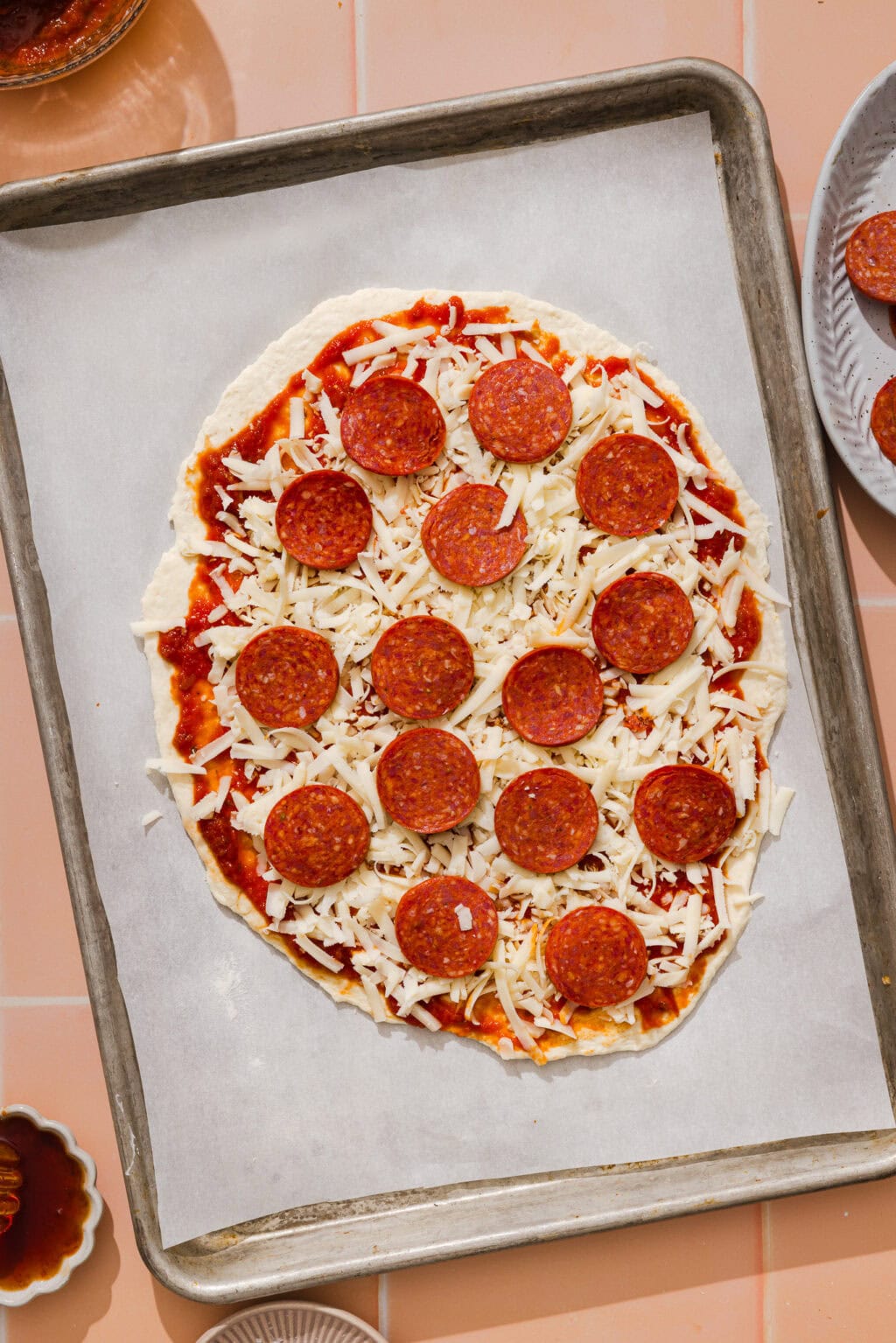 Uncooked pepperoni pizza with shredded cheese and tomato sauce on a baking sheet lined with parchment paper, surrounded by small bowls of ingredients on a peach-colored tiled surface.